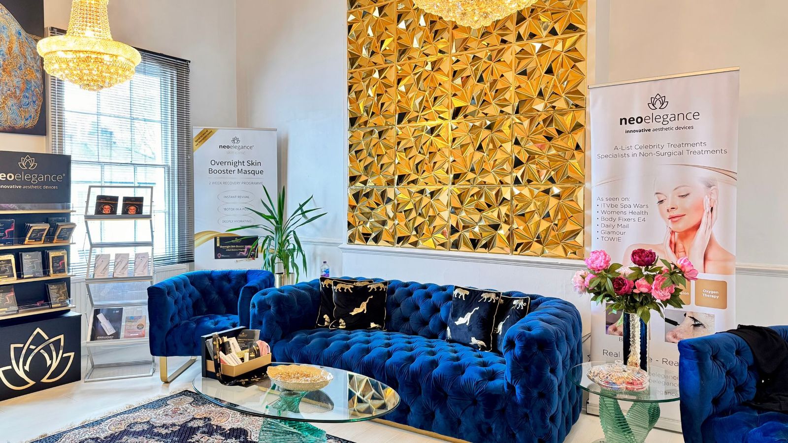 The client lounge at Neo Elegance Skin Clinic, Derby, with crystal chandeliers, a blue velvet sofa, a gold mirrored wall and fresh peonies